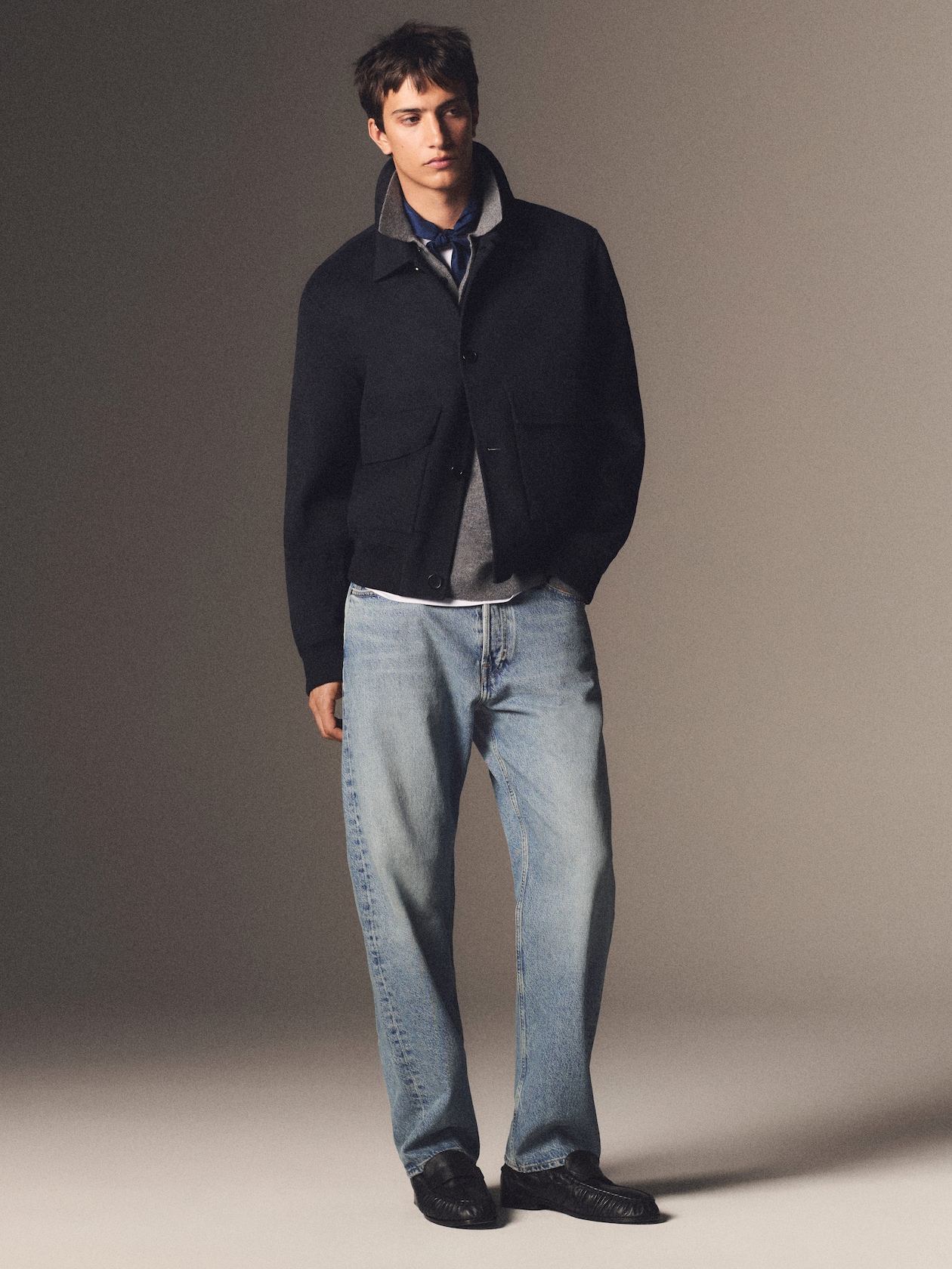A man wearing a dark jacket over a gray shirt with light blue jeans and black shoes, standing against a simple background.