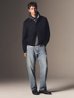 Placeholder for: A man wearing a dark jacket over a gray shirt with light blue jeans and black shoes, standing against a simple background.