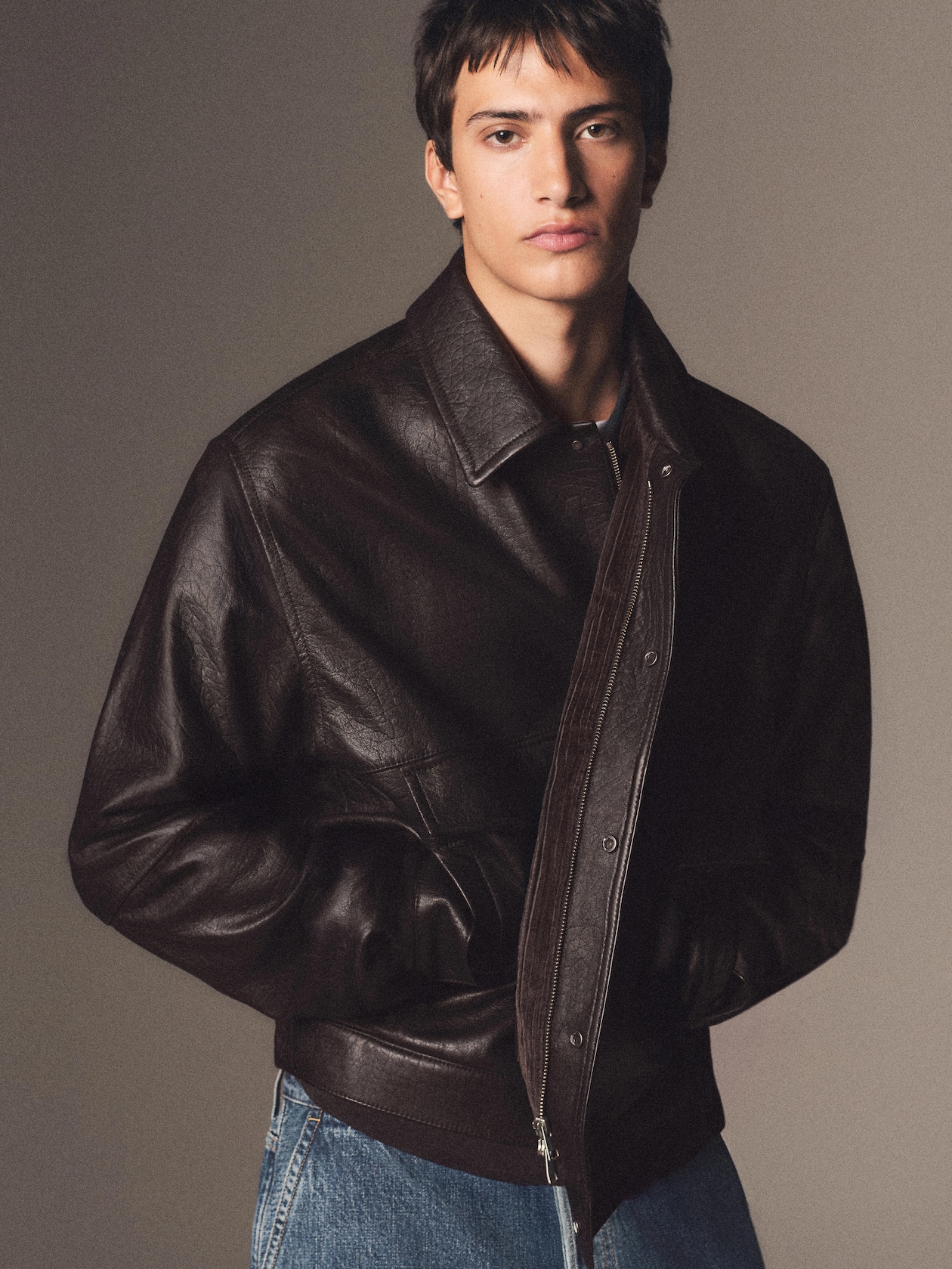 Young man in a dark brown leather jacket and blue jeans, hands in pockets, looking directly at the camera against a neutral background.