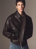Placeholder for: Young man in a dark brown leather jacket and blue jeans, hands in pockets, looking directly at the camera against a neutral background.