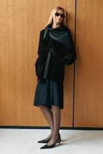 Placeholder for: Woman in sunglasses wearing a black coat and skirt, leaning against a wooden wall on beige carpet.
