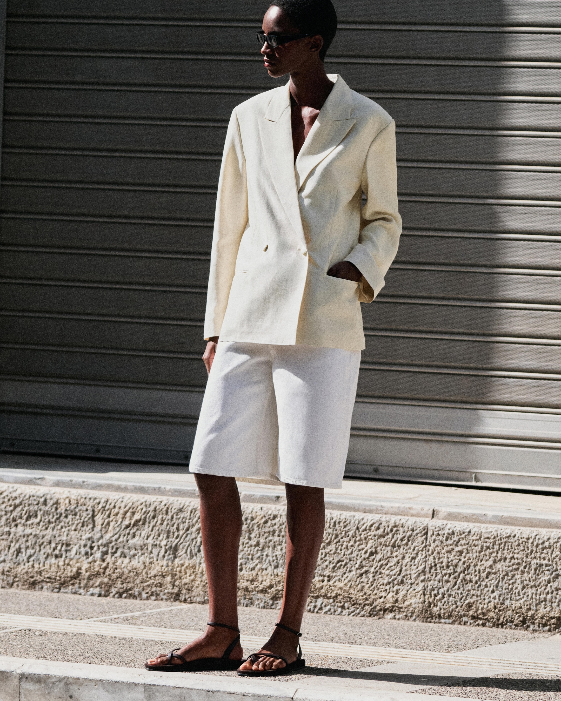 A person wearing a light beige blazer, white shorts, and sandals stands on a sunlit pavement in front of a closed shutter.
