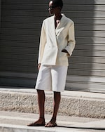 Placeholder for: A person wearing a light beige blazer, white shorts, and sandals stands on a sunlit pavement in front of a closed shutter.