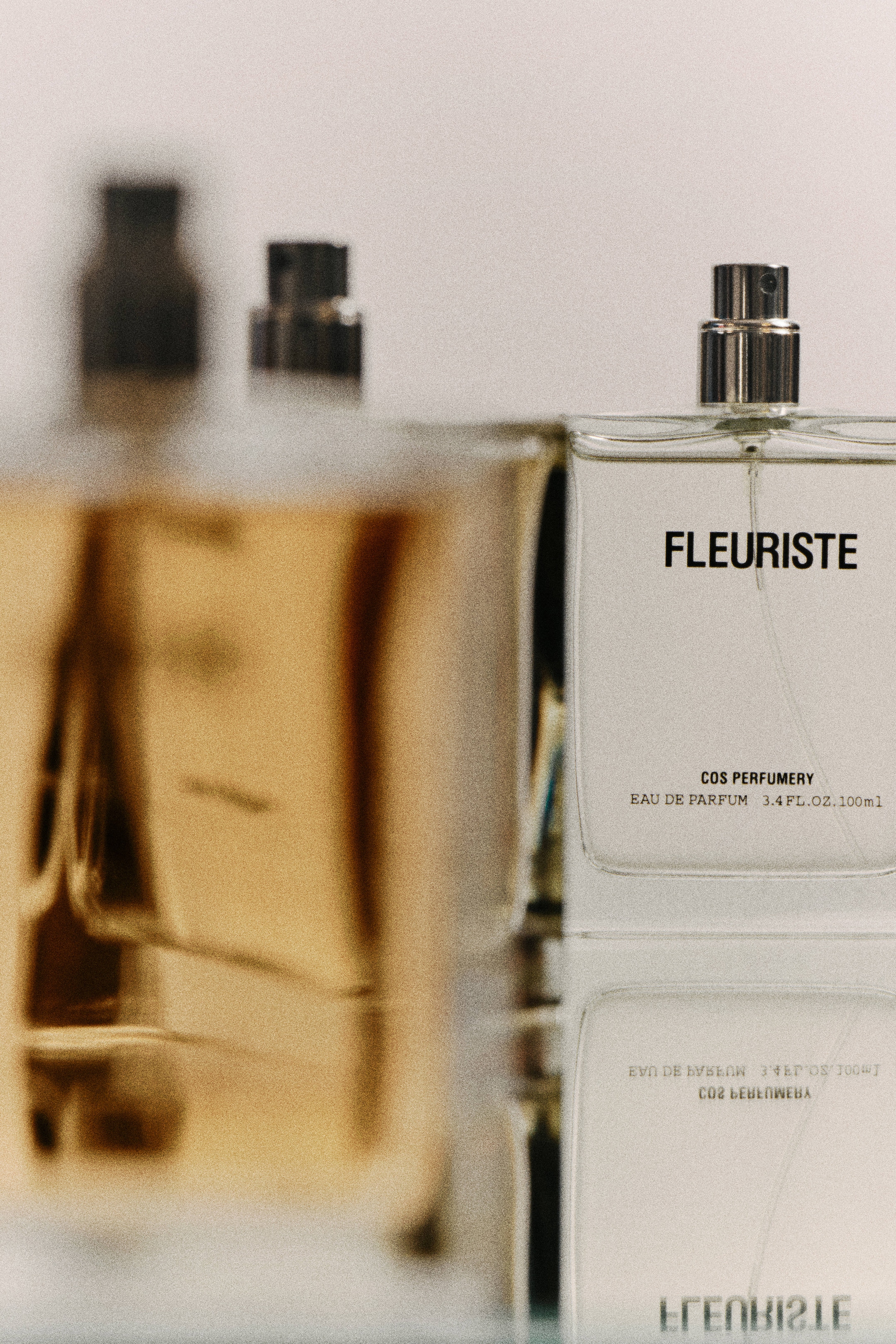 Close-up of perfume bottles on a reflective surface, focusing on a clear square bottle labeled "Fleuriste" by COS Perfumery, 100ml.
