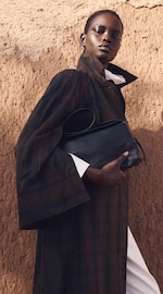 Placeholder for: Person in a long dark coat holding a black clutch against a textured brown wall.