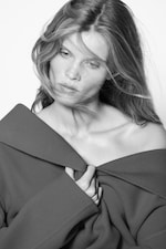 Placeholder for: Black and white portrait of a woman with long hair, wearing an off-shoulder coat, looking contemplatively to the side.