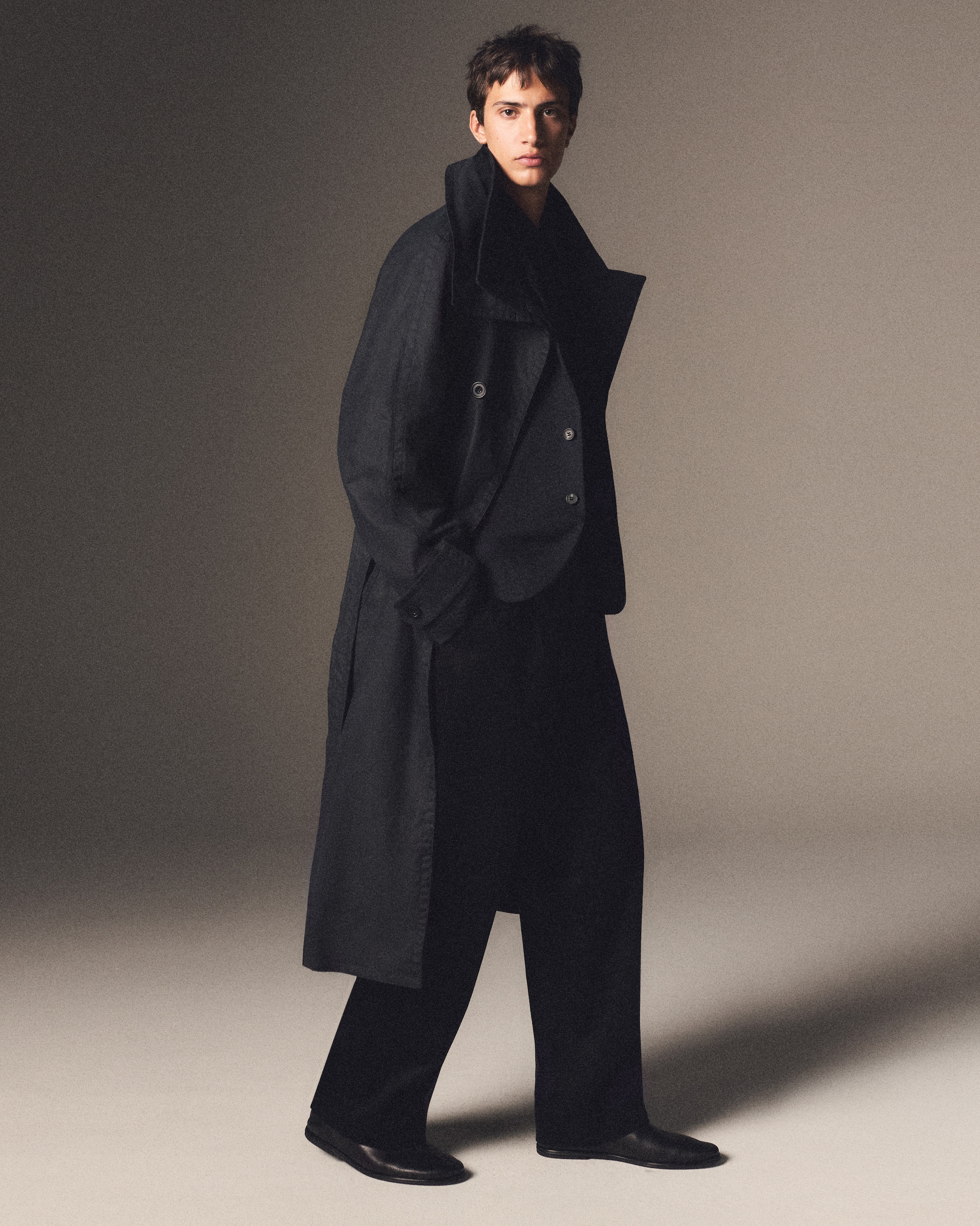 A person wearing a long, dark coat and wide trousers stands in a neutral-toned setting, looking confidently at the camera.