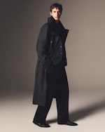 Placeholder for: A person wearing a long, dark coat and wide trousers stands in a neutral-toned setting, looking confidently at the camera.