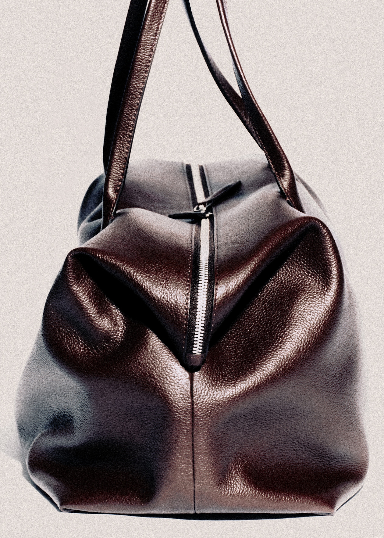 Brown leather duffel bag with textured surface, prominent zipper, and two handles, positioned upright against a plain background.
