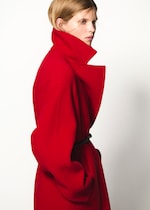 Placeholder for: Person in a bright red coat with a high collar, looking to the side against a plain white background.