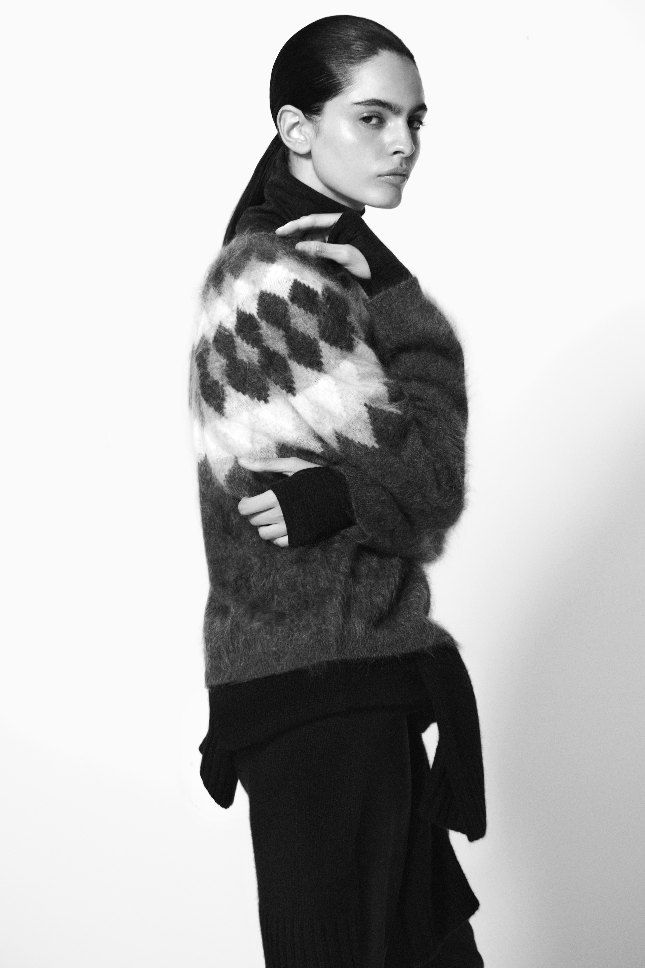 A person in a textured sweater with a diamond pattern poses against a plain background, looking to the side with a serious expression. Black and white image.