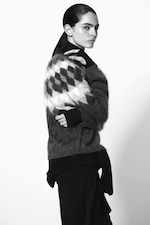 Placeholder for: A person in a textured sweater with a diamond pattern poses against a plain background, looking to the side with a serious expression. Black and white image.