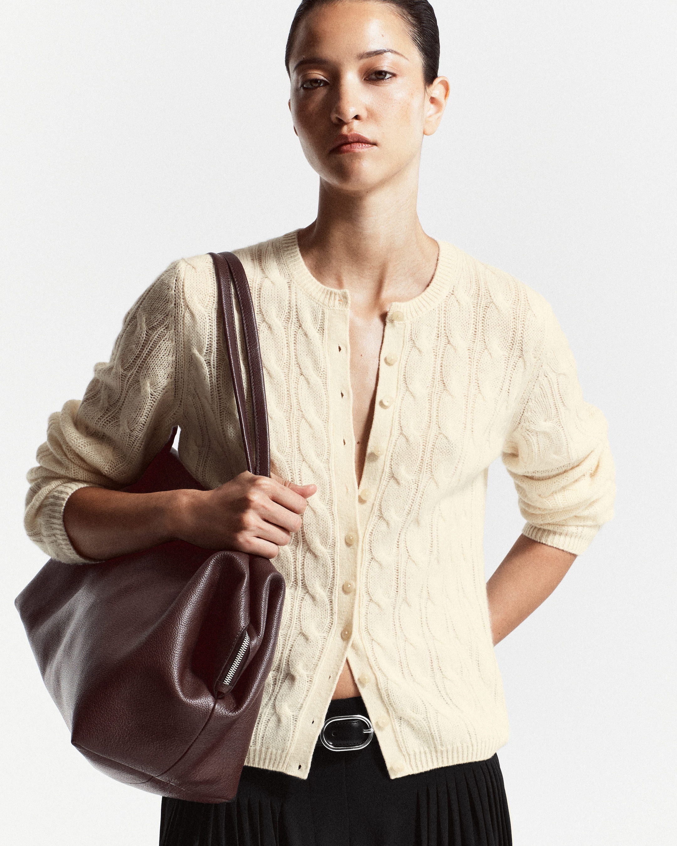A person wearing a cream cable knit cardigan and black skirt, holding a large brown leather bag, with a neutral expression.