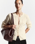 Placeholder for: A person wearing a cream cable knit cardigan and black skirt, holding a large brown leather bag, with a neutral expression.