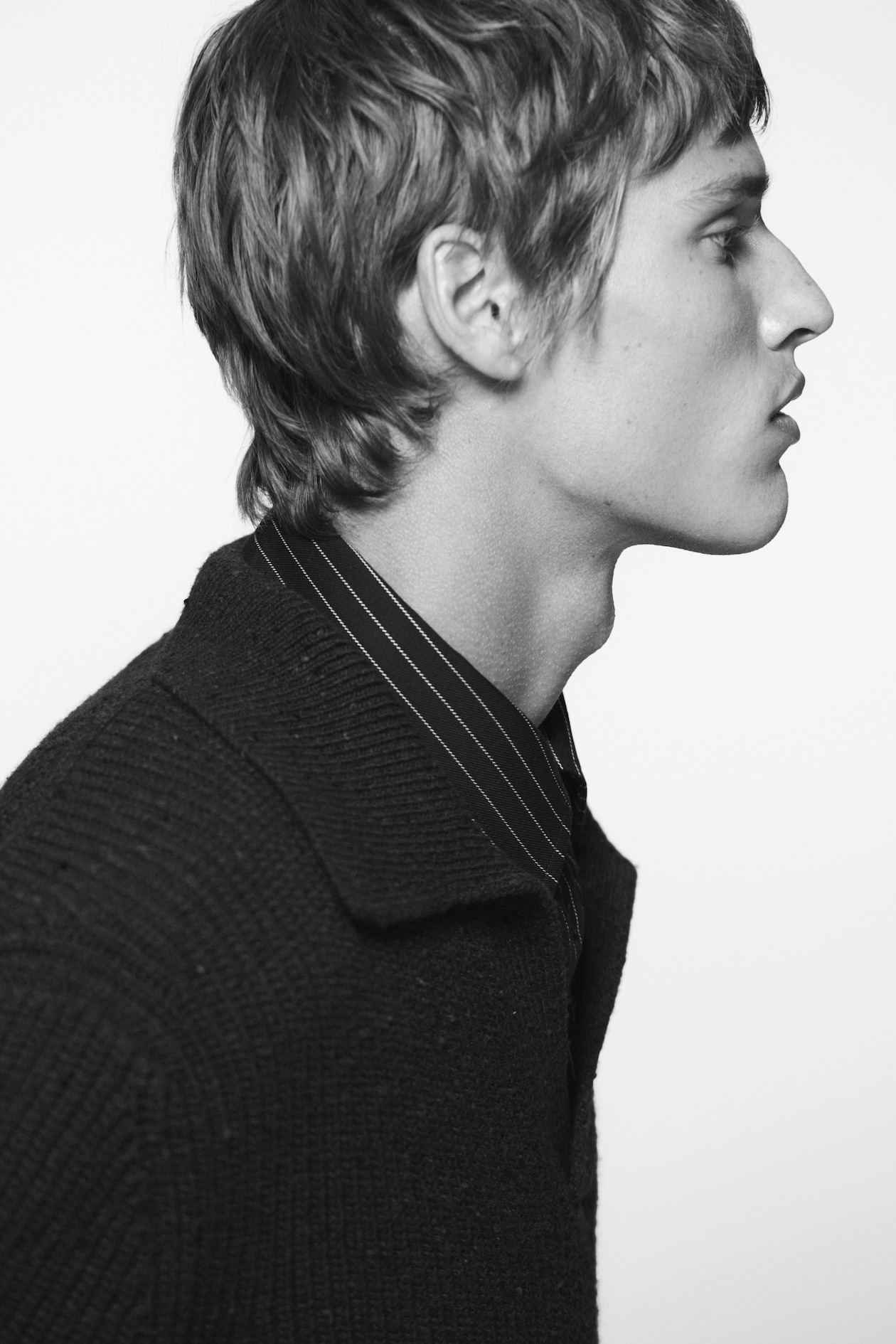 Profile of a person with short hair, wearing a dark sweater and striped shirt, looking to the right against a plain background.