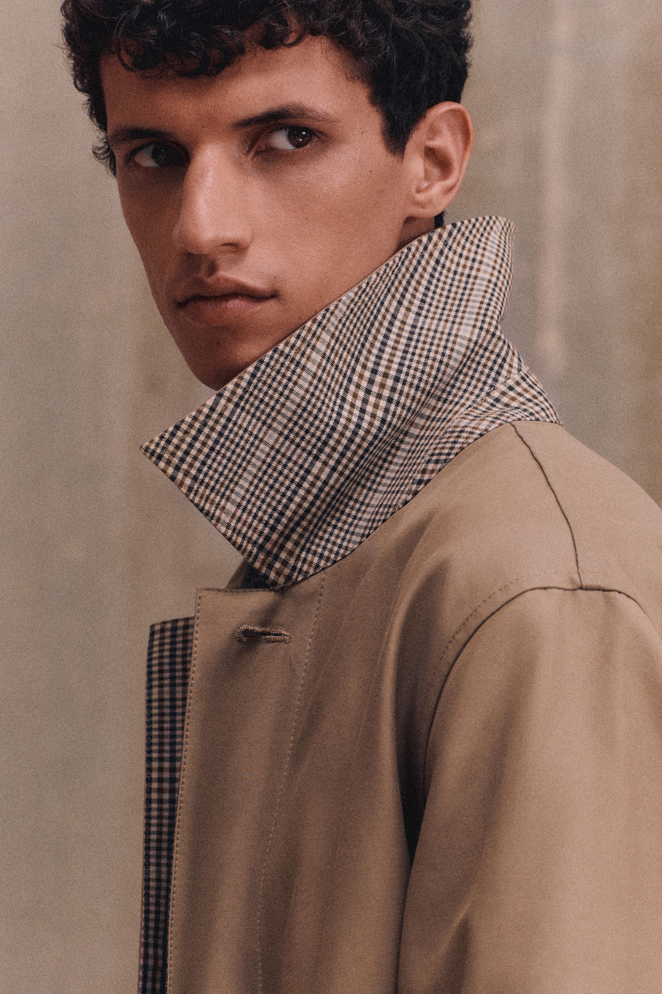 A person with curly hair wearing a beige trench coat with a checkered collar, looking to the side against a neutral background.