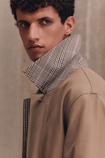 Placeholder for: A person with curly hair wearing a beige trench coat with a checkered collar, looking to the side against a neutral background.