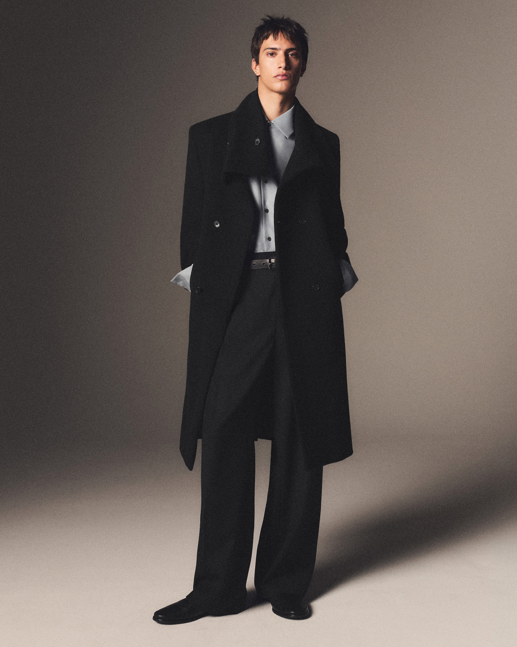 A person wearing a long black coat, light blue shirt, and black trousers stands against a neutral background, hands in pockets.