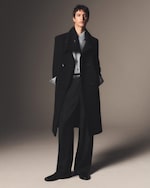Placeholder for: A person wearing a long black coat, light blue shirt, and black trousers stands against a neutral background, hands in pockets.
