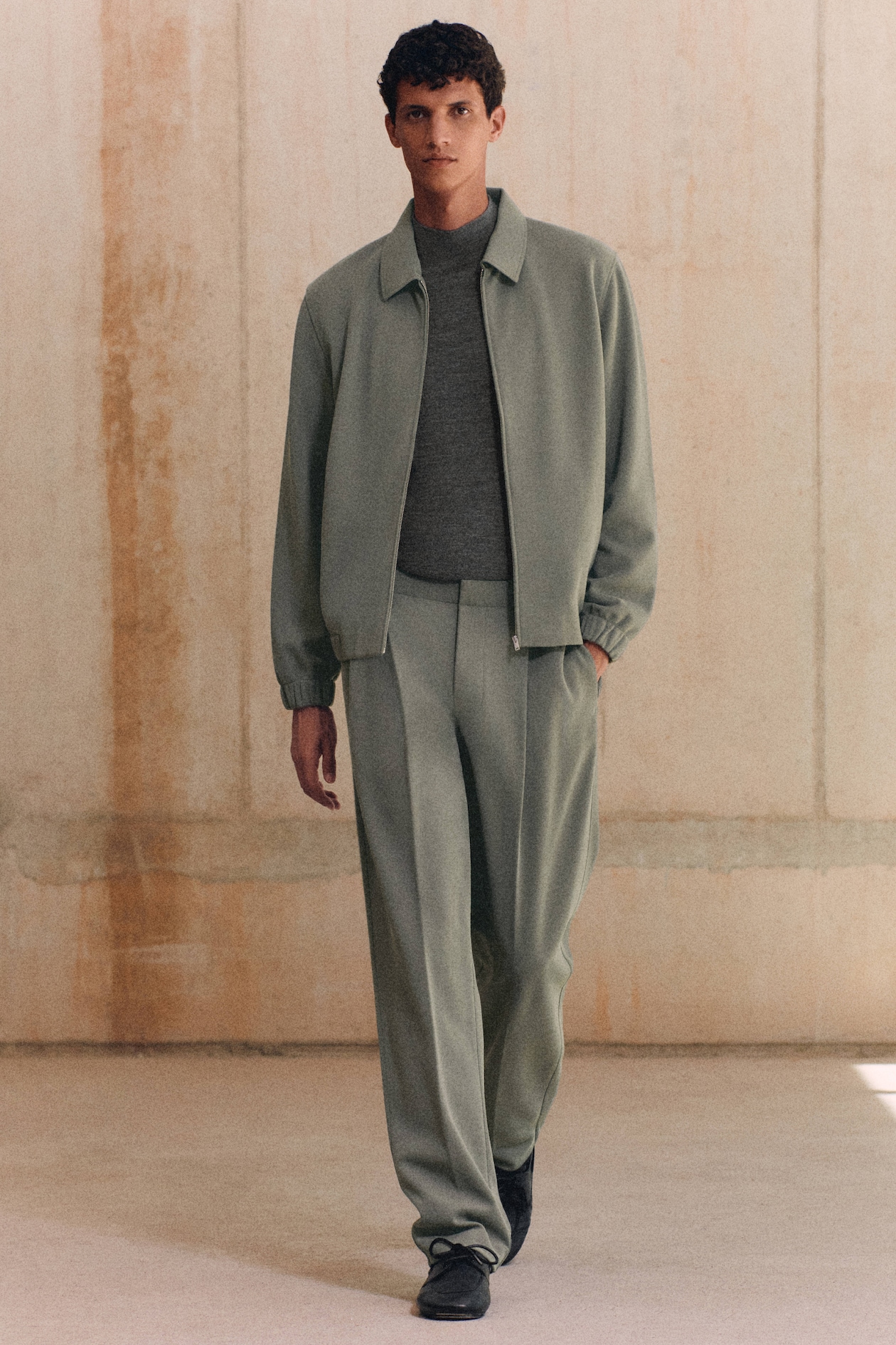A man in a green suit with a gray sweater walks in a minimalist indoor setting.