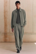 Placeholder for: A man in a green suit with a gray sweater walks in a minimalist indoor setting.