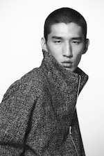 Placeholder for: Black and white portrait of a person with short hair, wearing a textured, high-collared jacket, looking directly at the camera.