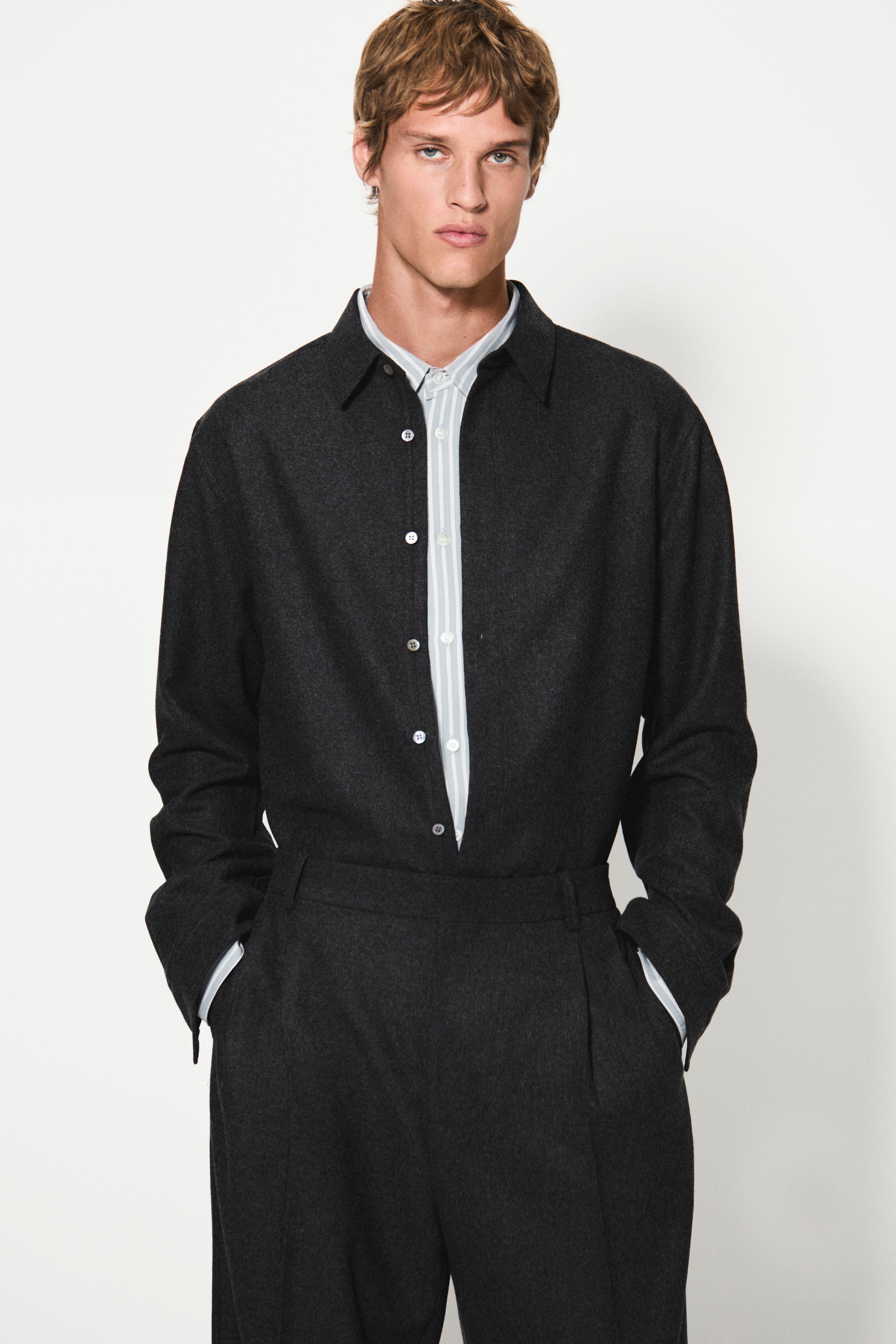 Person wearing a dark button-up shirt and matching pants, standing with hands in pockets against a plain white background.