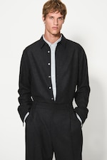 Placeholder for: Person wearing a dark button-up shirt and matching pants, standing with hands in pockets against a plain white background.