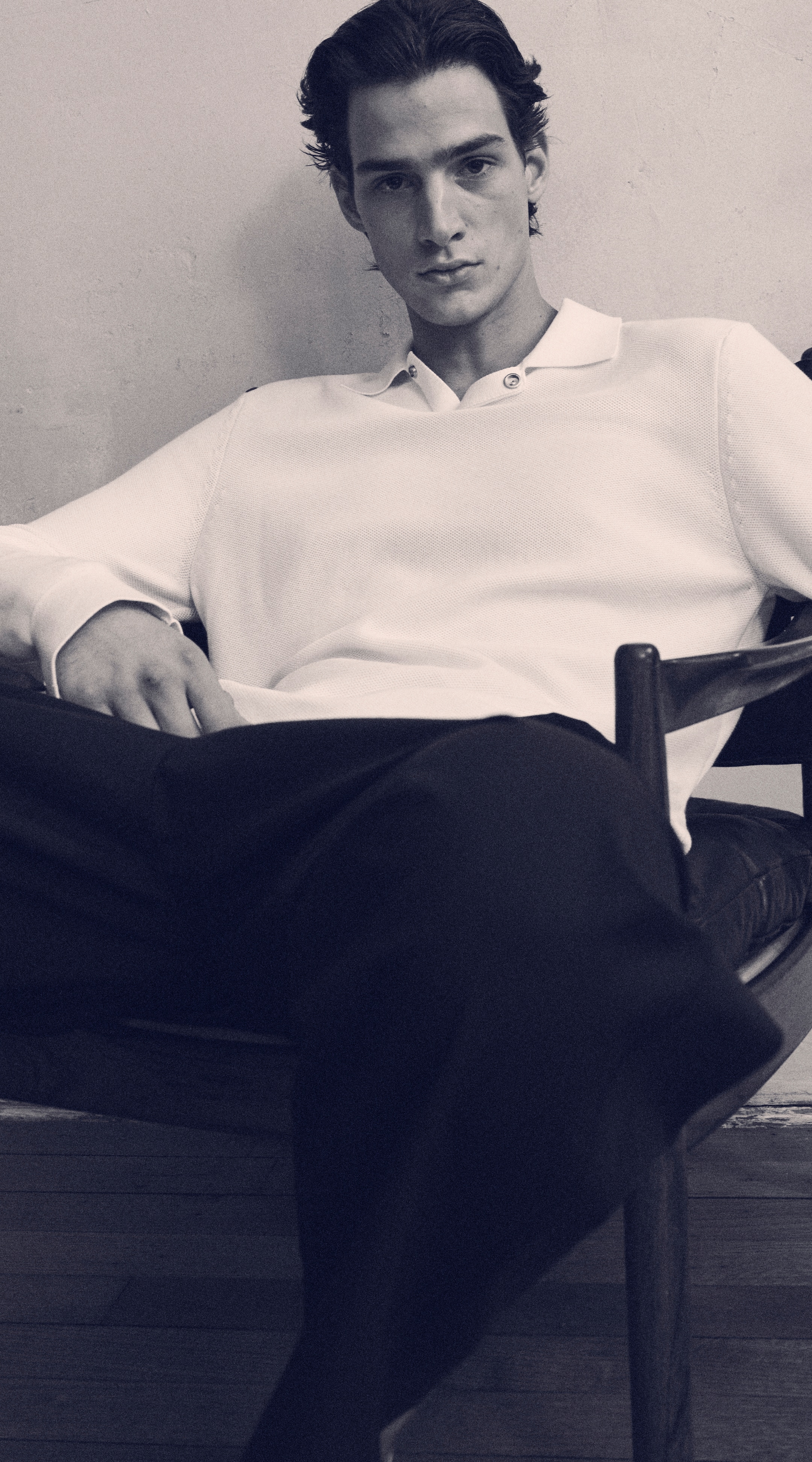 Black & white image of a man in a knitted white polo shirt & black trousers on, sitting down