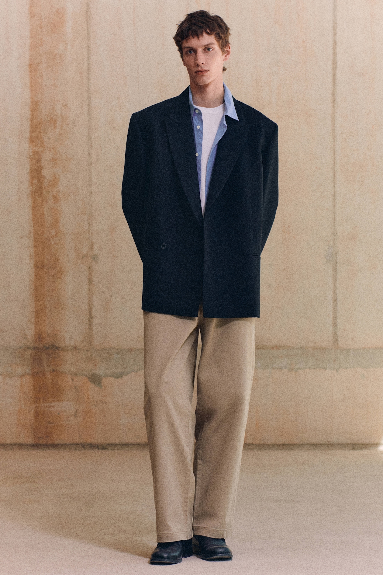 A person stands indoors wearing a black blazer, light blue shirt, beige pants, and black shoes against a plain wall background.