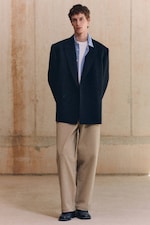 Placeholder for: A person stands indoors wearing a black blazer, light blue shirt, beige pants, and black shoes against a plain wall background.