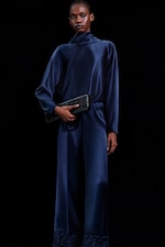 Placeholder for: Model in a dark blue satin outfit holding a black clutch against a black background.