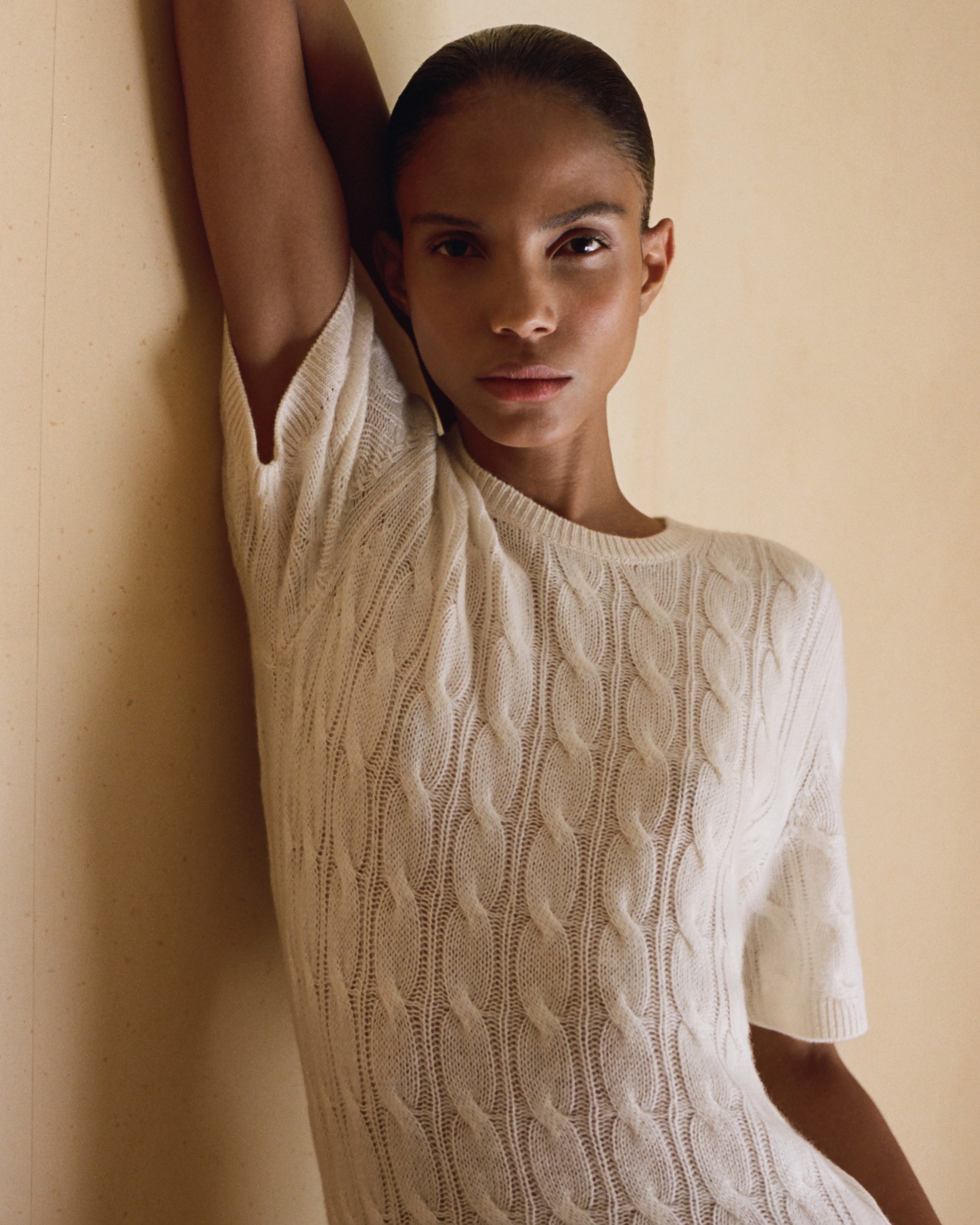 Person wearing a white cable-knit sweater, leaning against a wall with one arm raised, gazing directly at the camera with a neutral expression.