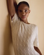 Placeholder for: Person wearing a white cable-knit sweater, leaning against a wall with one arm raised, gazing directly at the camera with a neutral expression.