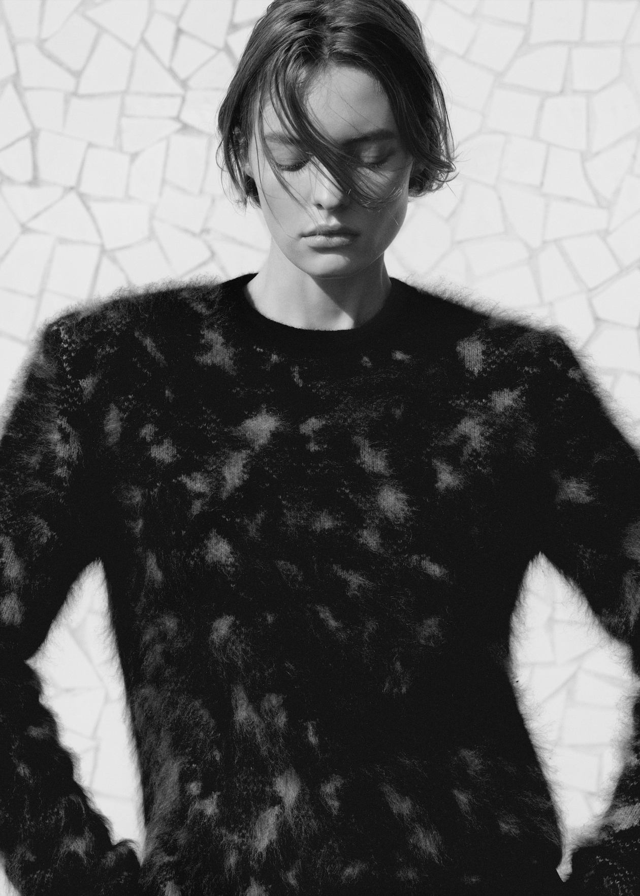 Black and white photo of a person with short hair wearing a textured, patterned sweater, standing against a tiled background.