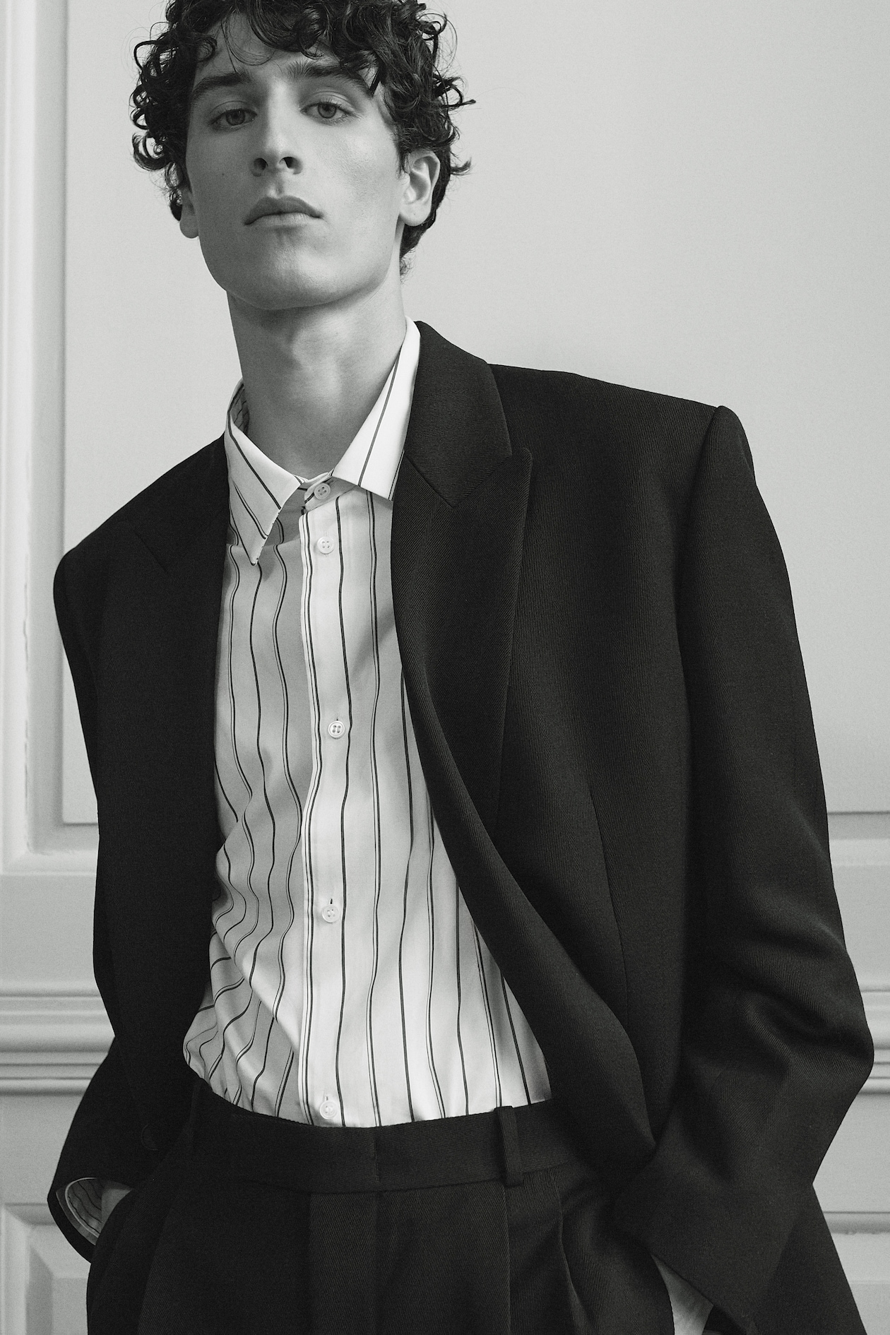 A young man with curly hair wearing a black suit and striped shirt, standing against a minimalist background.