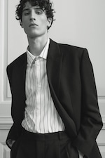 Placeholder for: A young man with curly hair wearing a black suit and striped shirt, standing against a minimalist background.