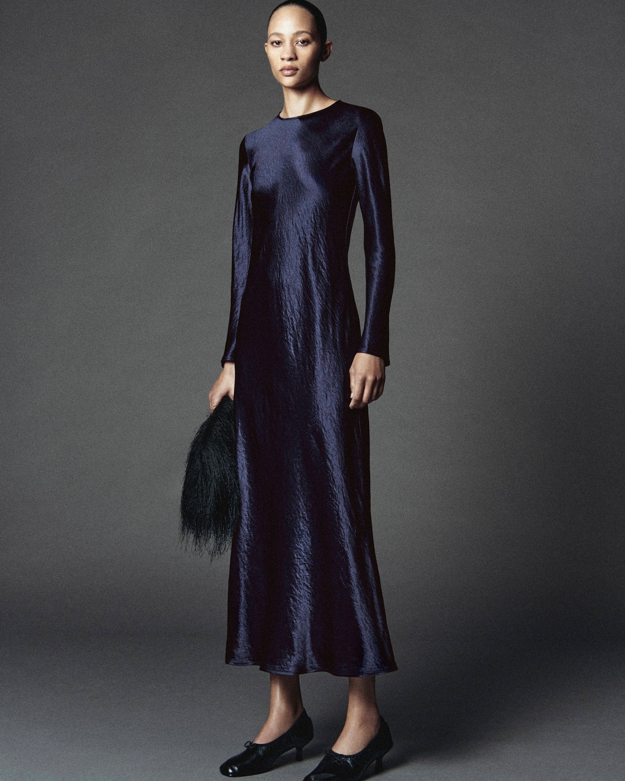 A woman poses in a dark, long-sleeved, satin dress with a textured look, holding a black accessory against a gray background.