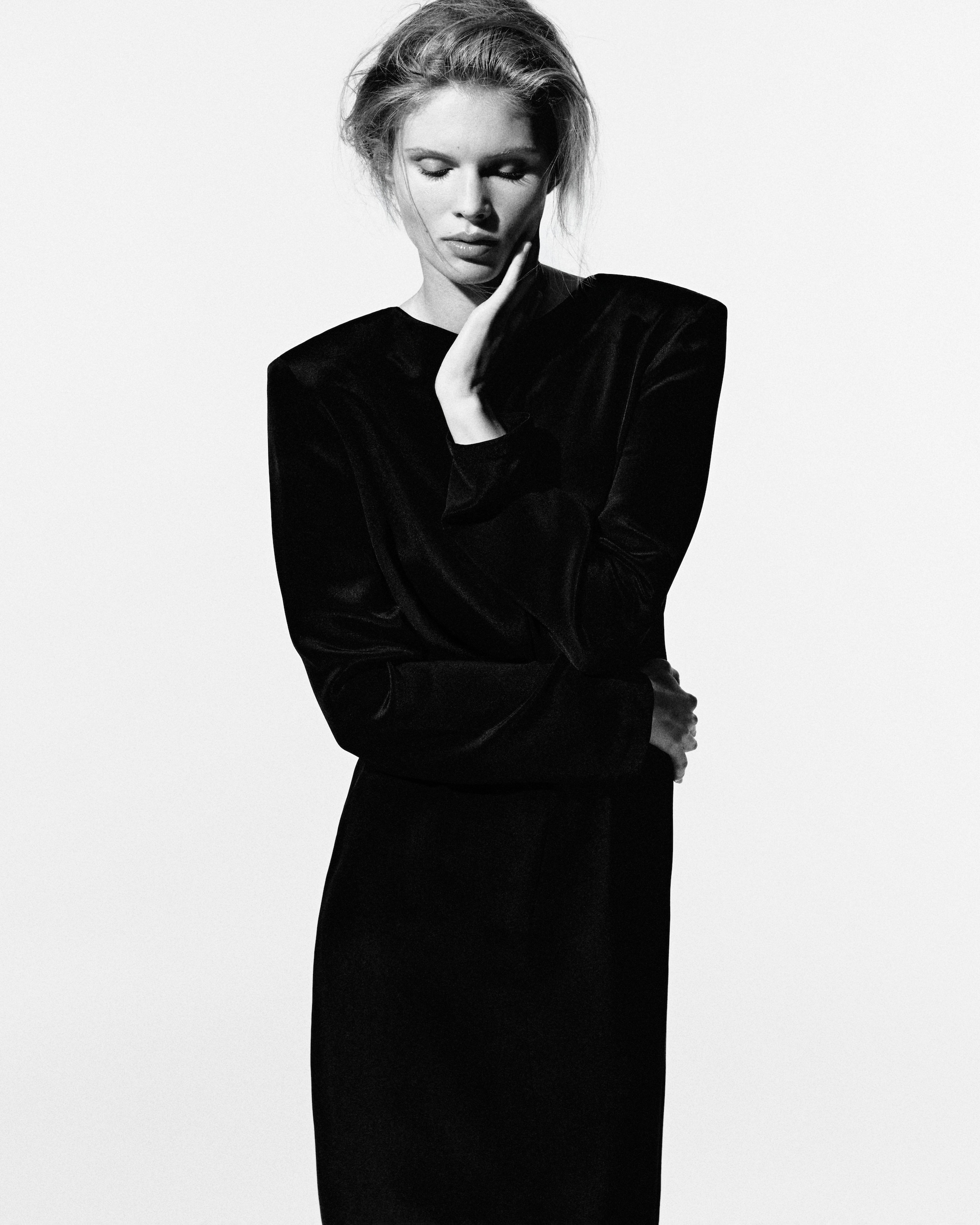 Black and white portrait of a person in a long-sleeved black dress, eyes closed, resting hand on chin, with a contemplative expression.