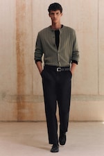 Placeholder for: A person stands confidently wearing a green cardigan, black pants, and a black belt, with hands in pockets, set against a neutral background.