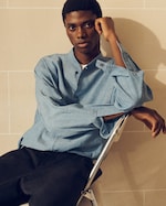 Placeholder for: A person in a light blue shirt sits on a chair with one arm resting on the knee and the other elbow propped on the backrest, against a tiled wall.
