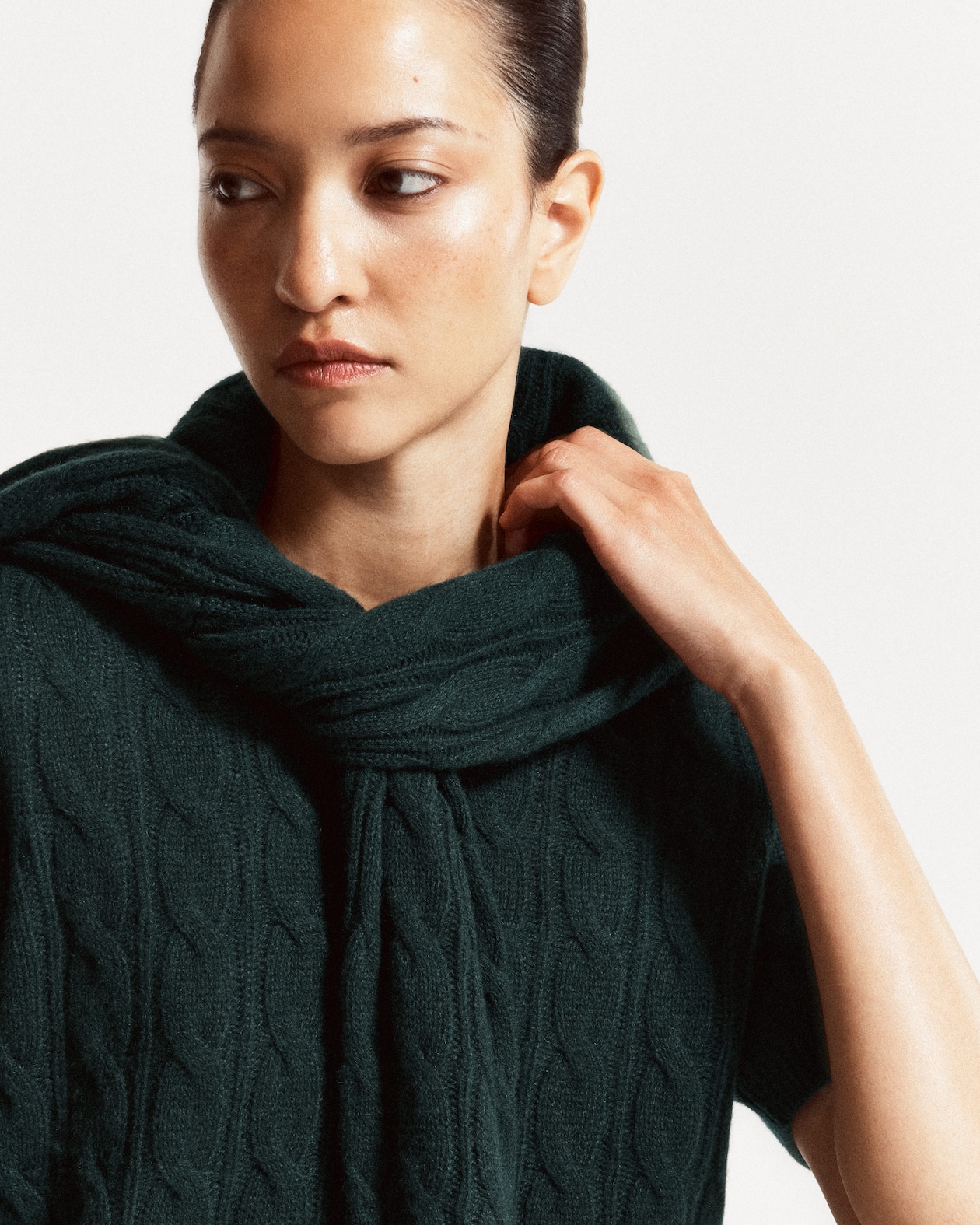 A woman gazes to the side, wearing a dark green, textured knitted garment with a cozy wrap and short sleeves, set against a light background.