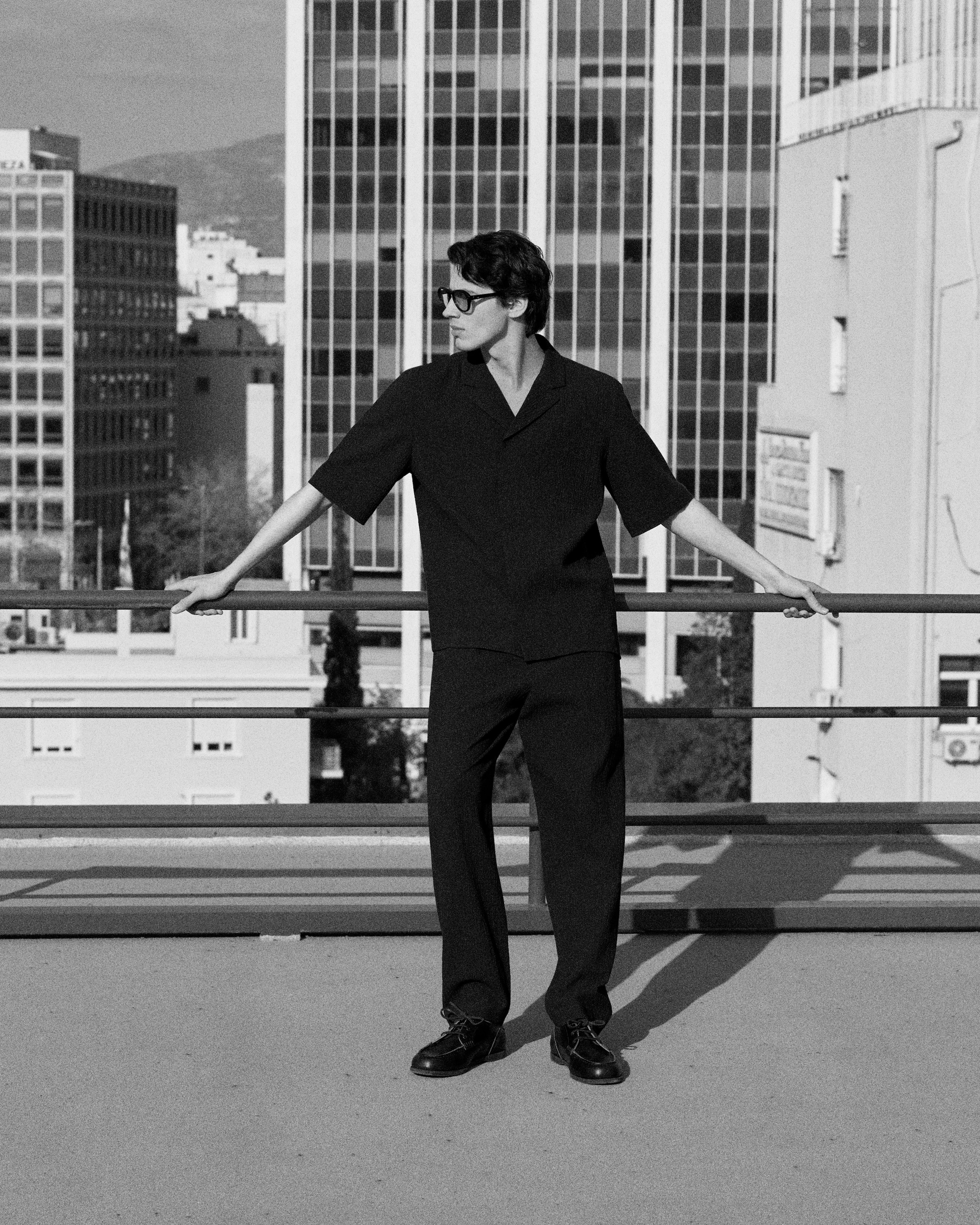 A person wearing sunglasses stands confidently on a rooftop, leaning on a railing, with tall buildings in the background. Black and white image.