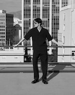 Placeholder for: A person wearing sunglasses stands confidently on a rooftop, leaning on a railing, with tall buildings in the background. Black and white image.