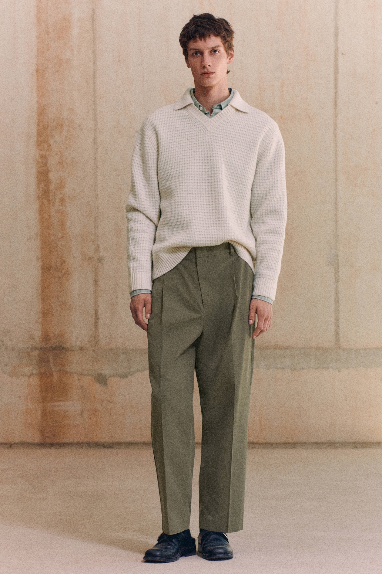 A person wearing a white textured sweater over a green shirt with olive trousers and black shoes, standing against a neutral, textured background.