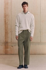 Placeholder for: A person wearing a white textured sweater over a green shirt with olive trousers and black shoes, standing against a neutral, textured background.