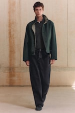 Placeholder for: A person wearing a green jacket over a dark polo shirt and black jeans walks indoors on a light floor with a neutral wall background.