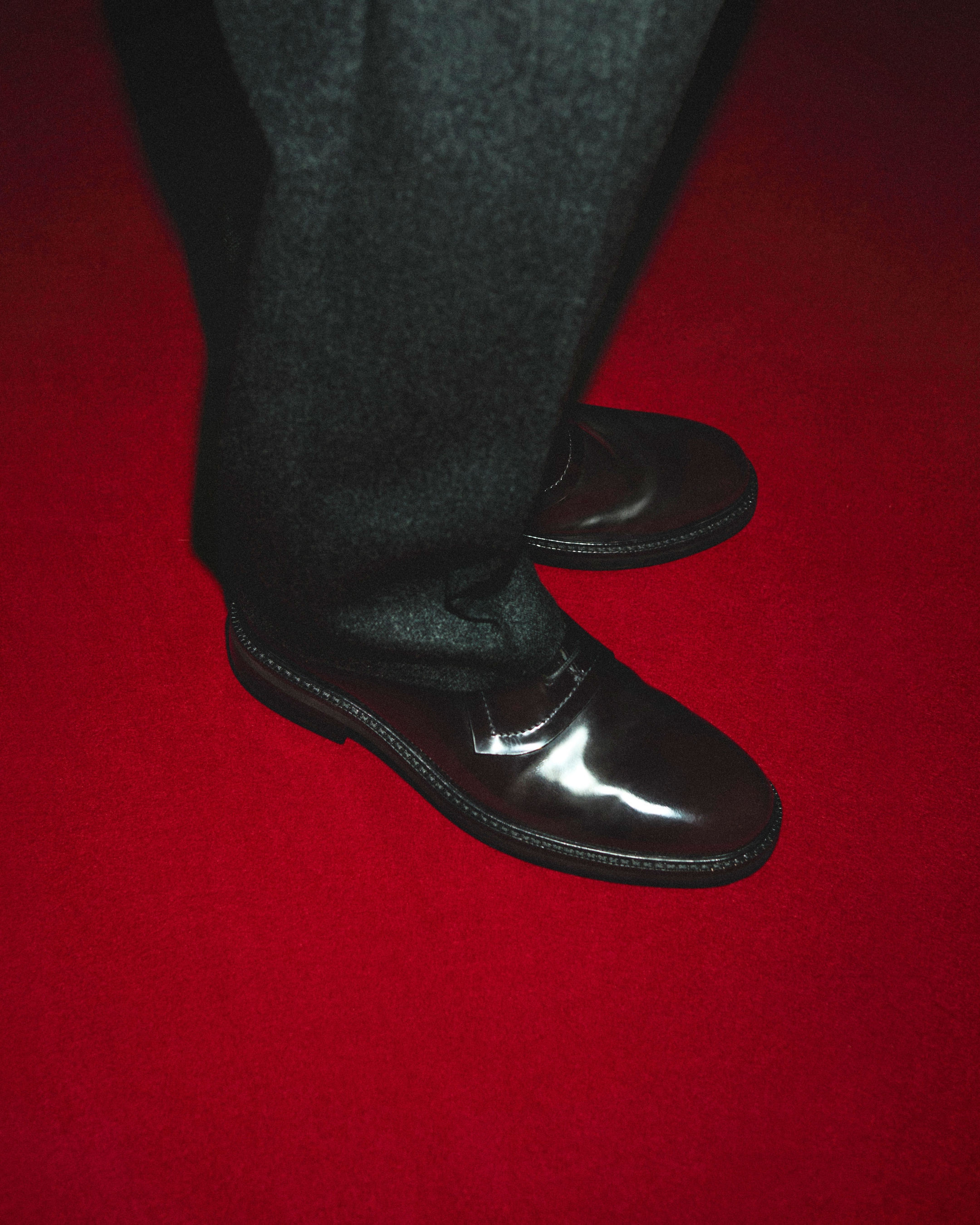 Man in dark pants and shiny black dress shoes standing on a red carpet.
