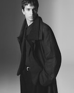 Placeholder for: Black and white portrait of a young person in a dark trench coat, hands in pockets, gazing into the distance with a serious expression.