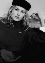 Placeholder for: Black and white portrait of a person with long hair wearing a dark turtleneck sweater and a black hat, leaning back with an arm bent.